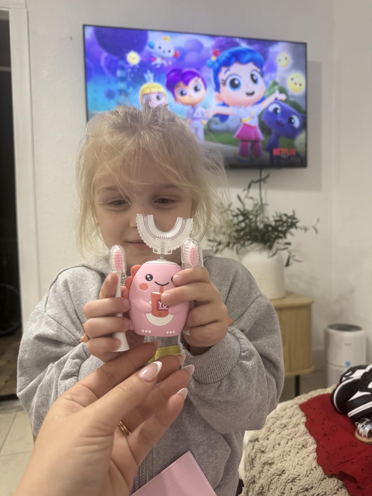 Child holding a pink and white toy with a TV in the background displaying animated characters.